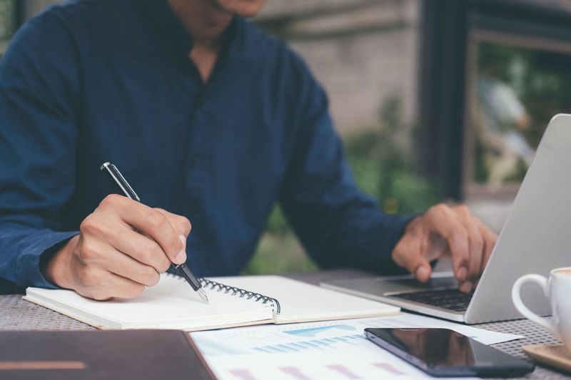 Closeup of someone's workspace with them working at a laptop and taking notes with a pen and notepad